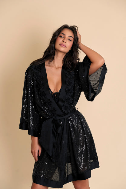 A woman modeling a black Stella Sequin Kimono Robe with sequin fabric and floral neckline detail, paired with a black bodysuit.