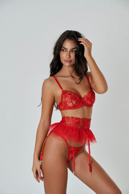 A woman wearing a red lingerie set with floral embroidery, including a bralette, Brazilian panty, and garter, with her hands behind her head.